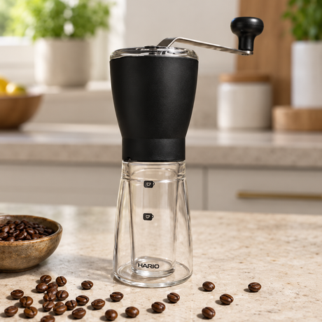 Tiamo manual coffee grinder with a black body and clear glass lower chamber, set on a kitchen countertop with scattered coffee beans.