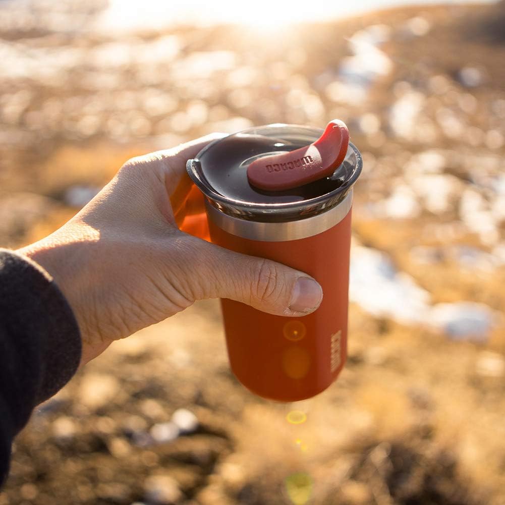Hand holding a red insulated mug with a black lid against a blurred natural background