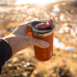 Hand holding a red insulated mug with a black lid against a blurred natural background