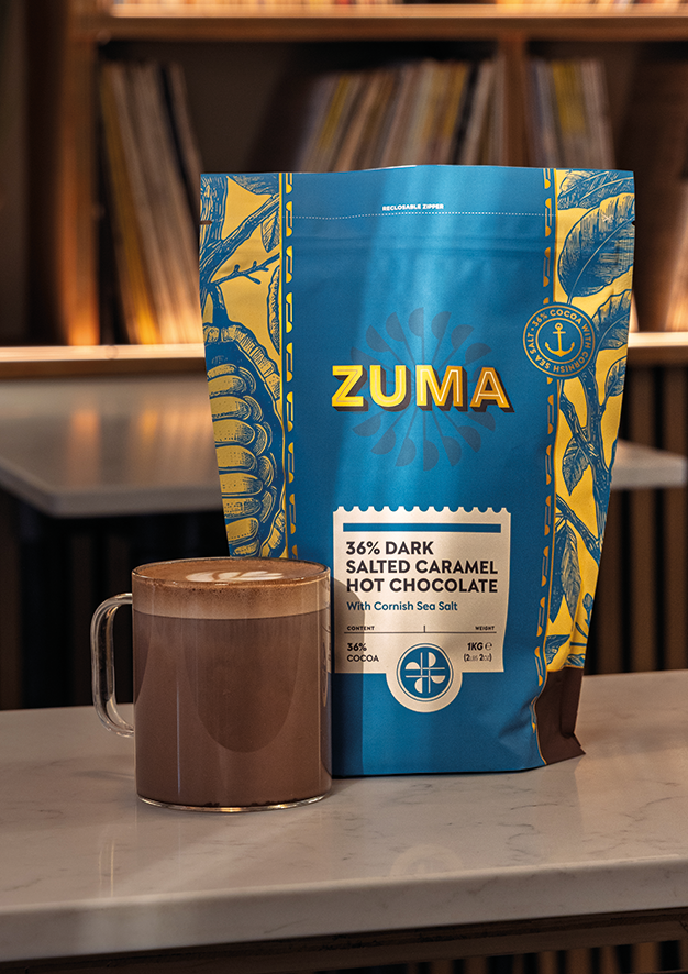 Zuma 60% Dark Salted Caramel Hot Chocolate package with a mug of hot chocolate on a table.