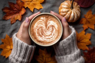 Person holding a cup of coffee with latte art surrounded by autumn leaves and a small pumpkin.
