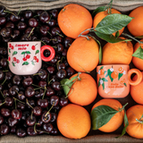 Bialetti Cherry and Orange porcelain mugs displayed among cherries and oranges