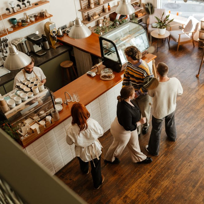 People waiting in line at a coffee shop with a barista preparing drinks.