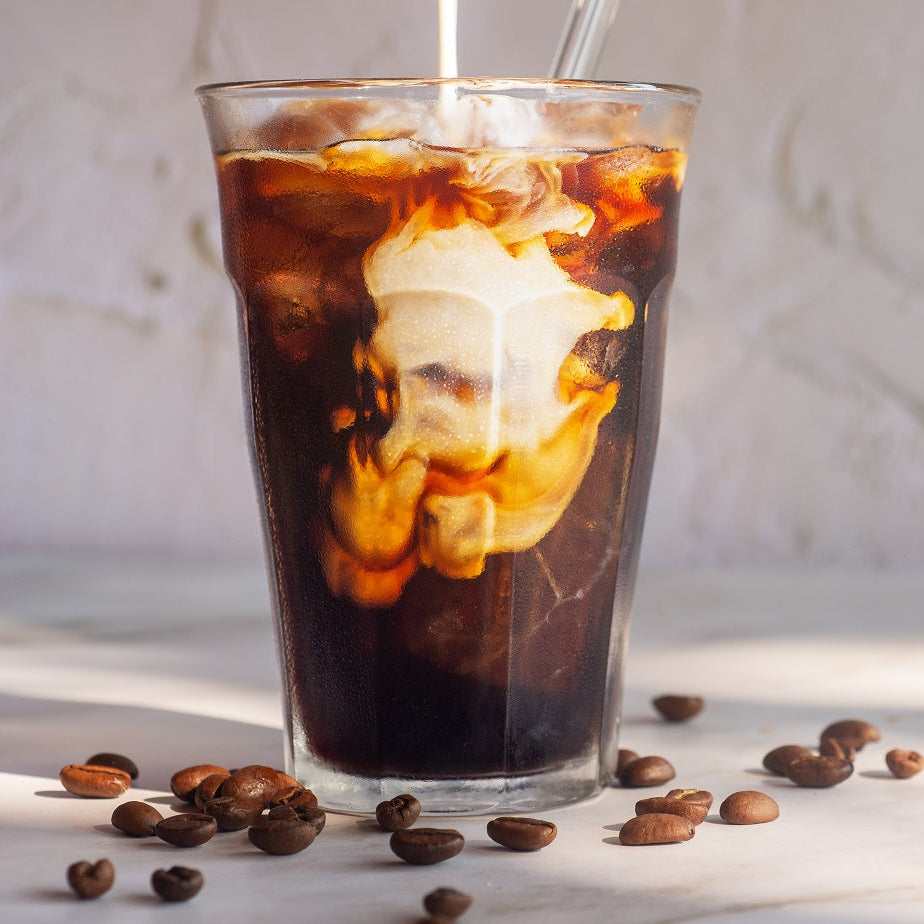 Glass of iced coffee with ice cubes and a straw, surrounded by coffee beans on a light surface.